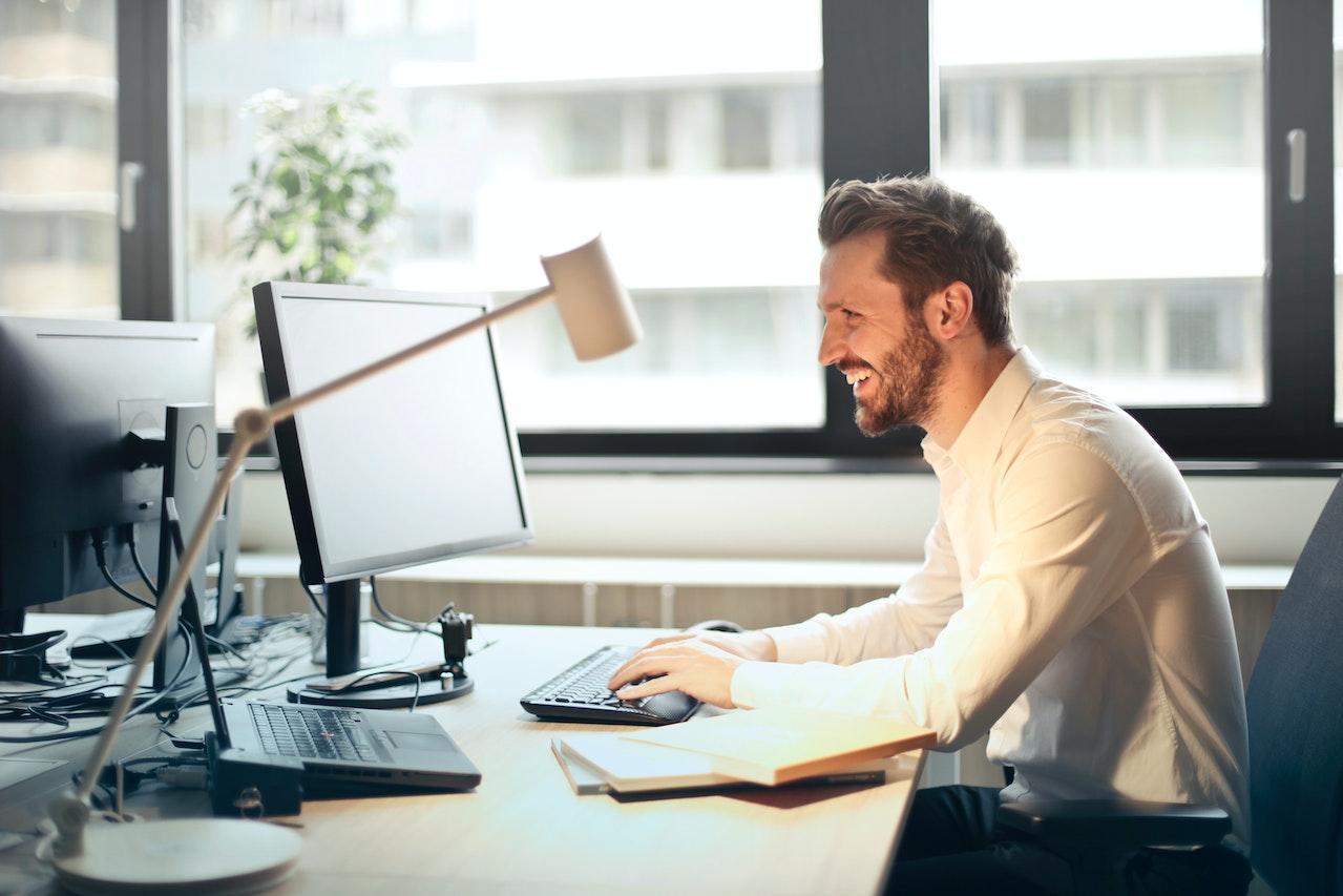 A man with a beard, wearing a white shirt, sits at a desk with two computer monitors, a keyboard, a mouse, a desk lamp, and a notebook, with a window and plant visible behind him.