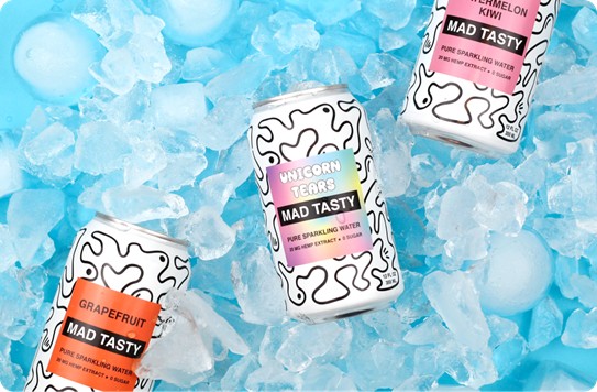 Three Mad Tasty sparkling water cans in Grapefruit, Unicorn, and Watermelon flavors arranged on a bed of blue ice cubes. The cans feature a distinctive white and black squiggly pattern design with colorful flavor labels.