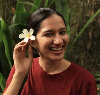 A woman with dark hair, wearing a red shirt, holds a white flower with five petals near her face, smiling and looking to the side. Green leaves and a tree trunk are visible in the background.